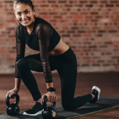Uśmiechnięta kobieta w stroju sportowym klęczy na macie z odważnikami kettlebell.
