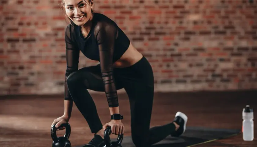 Uśmiechnięta kobieta w stroju sportowym klęczy na macie z odważnikami kettlebell.