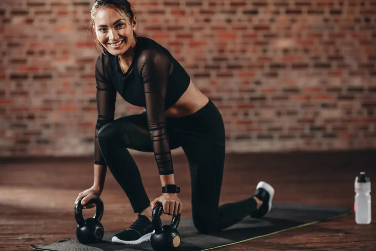 Uśmiechnięta kobieta w stroju sportowym klęczy na macie z odważnikami kettlebell.